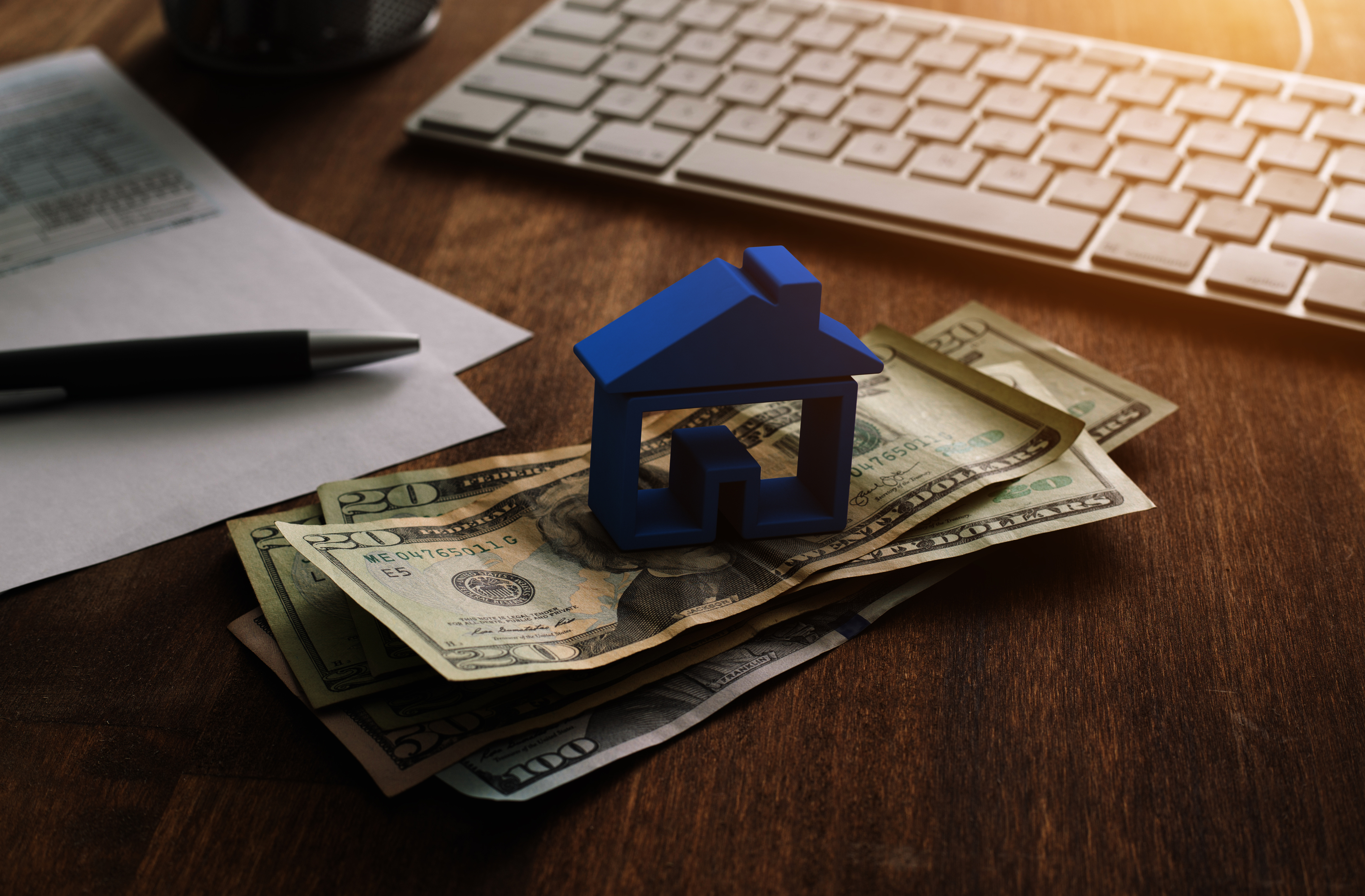 A blue wooden toy house over a pile of $20 bills aside a keyboard, paper, and pen