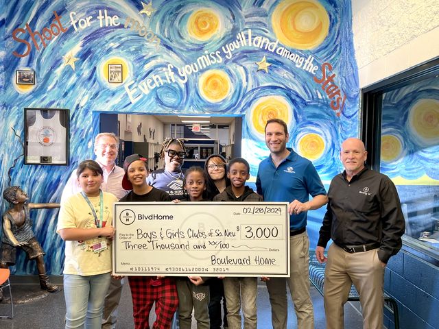 front view of a BlvdHome representative with the leaders and children of the Boys and Girls Club of Southern Nevada, holding a large check for three thousand dollars in a room painted blue and yellow