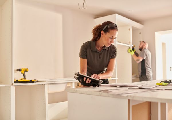 A woman reviewing remodeling plans while a man builds in the background 