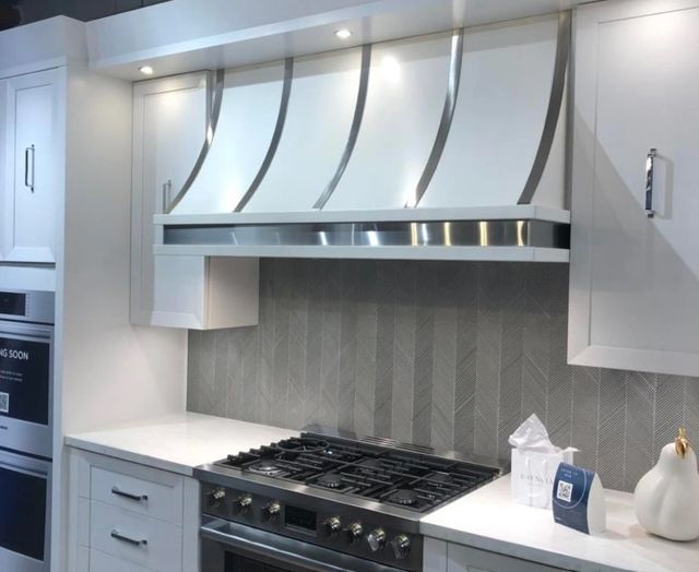 front view of a modern aire custom range hood above a stainless steel range with white cabinetry surrounding 