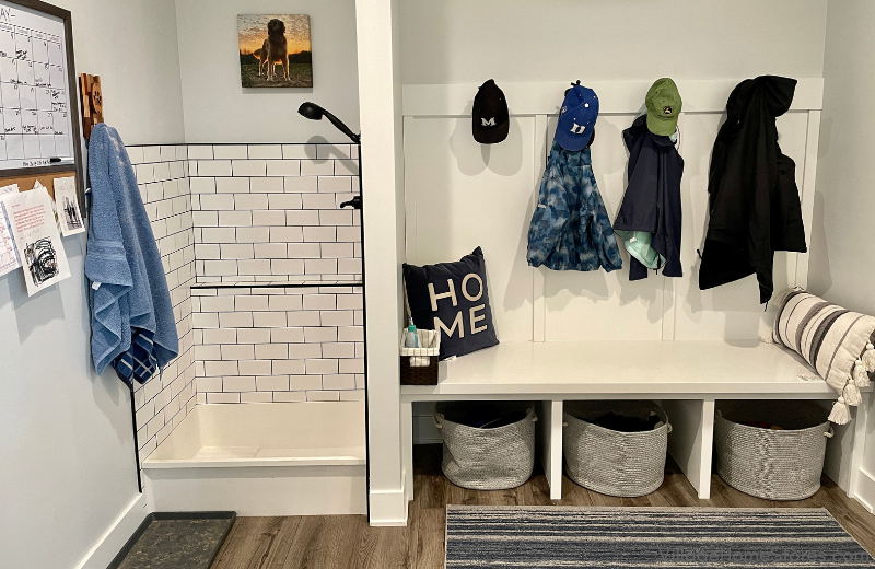 Image of painted white mudroom bench with coat hooks on right and white tiled dogwash shower on left. Blue towel hangs on a wall hook on left side of image.