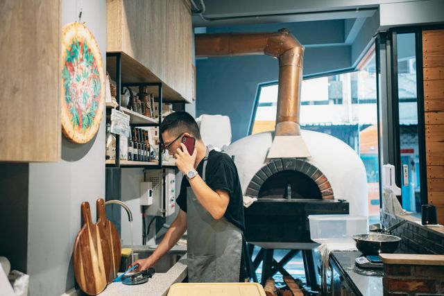 Front view of a small galley kitchen with a large pizza oven, properly vented, and a man standing on his phone by the sink.