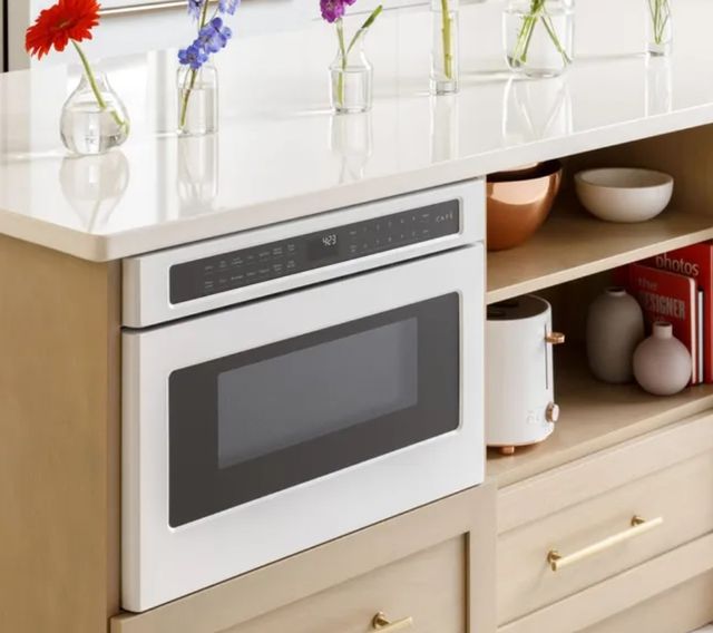 front left angled view of a white microwave drawer built into beige cabinets with shelving around and flower vases on the countertop