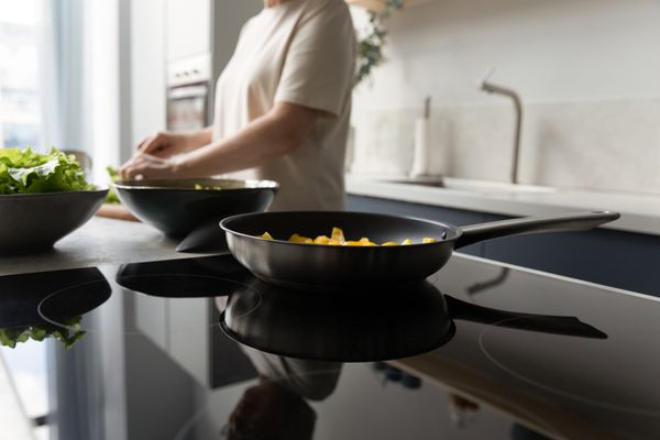 A close-up of a pan on a cooktop with a woman preparing food in the background 