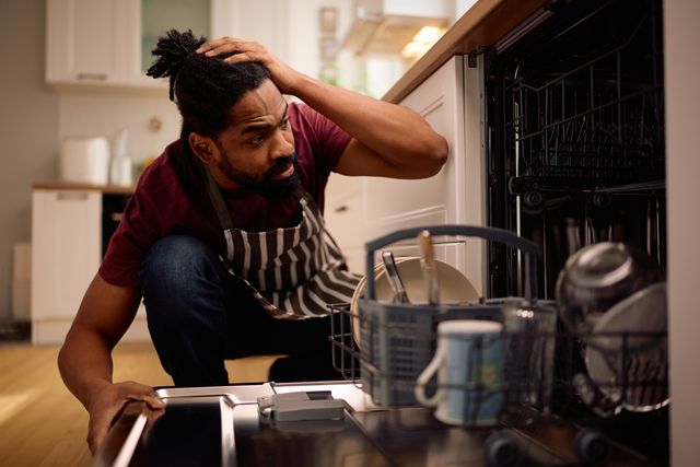 Front view of a man looking confused into his dishwasher