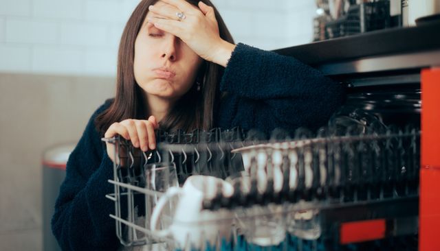 Front view of a woman looking defeated in front of her dishwasher