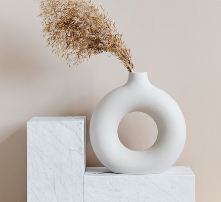 A round, donut-like vase sitting on a stone display platform with dried florals in it." title="A round, donut-like vase sitting on a stone display platform with dried florals in it