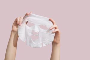 A person holding up a face mask sheet against a pink background 