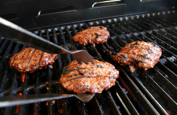 Four fully-cooked burgers being taken off the grill with metal tongs.