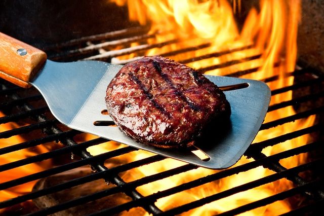 close view of a burger being grilled on a large flame, with a spatula under it about to flip