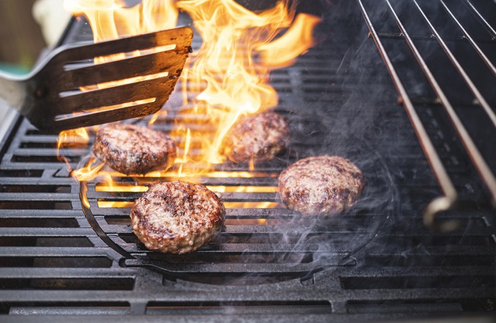 Four burger patties on a fiery grill with visible flames. 