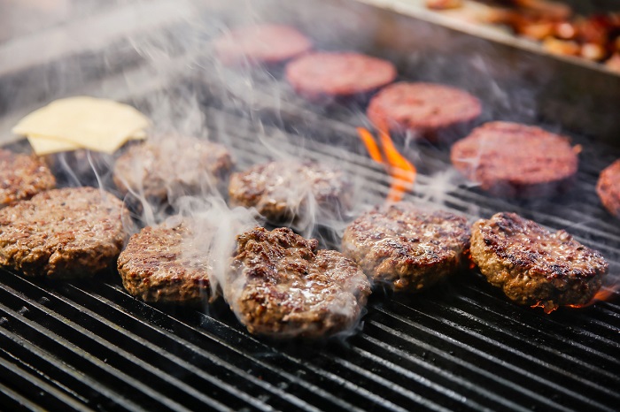 Rows of burgers on a fiery grill with visible smoke.