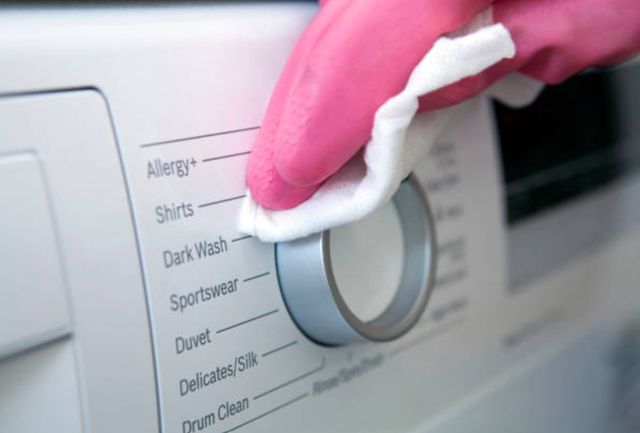 closeup view of a hand wearing a pink glove using a white cloth to wipe the control knobs of a washing machine