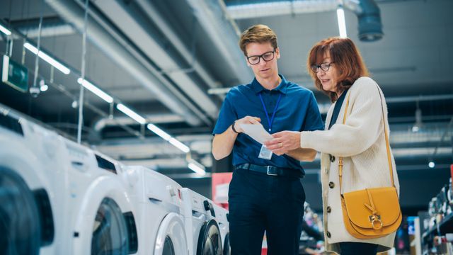 Front view of a salesman reviewing appliance rebates with a woman in a washing machine aisle of an appliance store