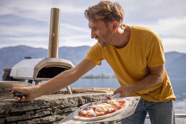 Front view of a man holding a pizza in front of an outdoor pizza oven