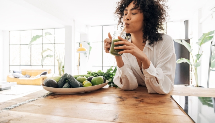 A woman happily sips a green smoothie in a brightly lit room.