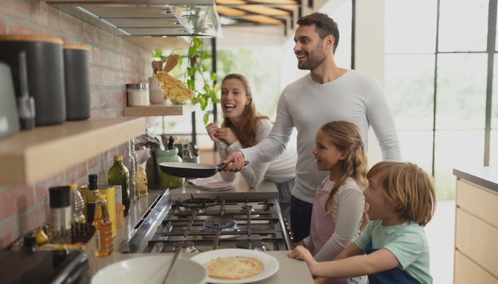 A father flips a pancake at the stove in front of his cheering wife and two children.