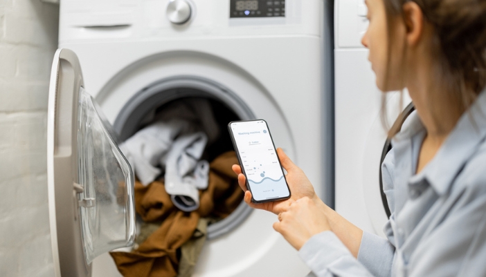 A woman sits outside an open washing machine and plans a load with her mobile phone.