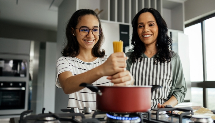 A young woman and her mom put pasta in a pot to cook together.