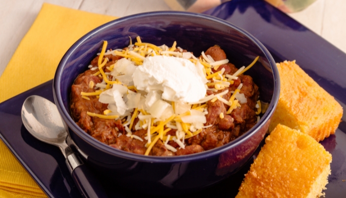 A blue bowl full of chili next to slices of corn bread.