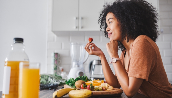 A woman eats a strawberry in her kitchen with a plate of fruit in front of her.