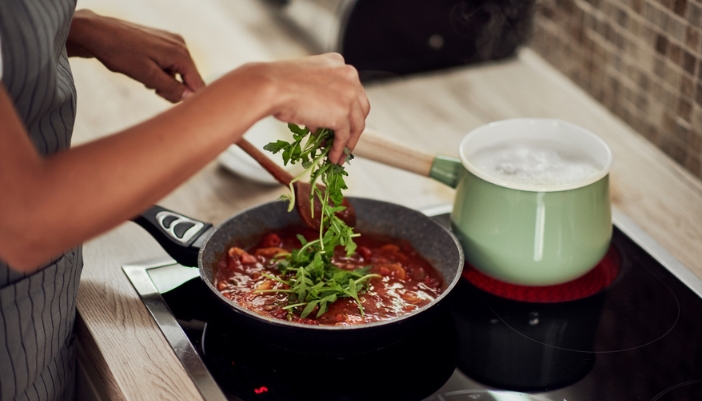 A pair of hands adds greens to her pasta sauce while pasta boils.