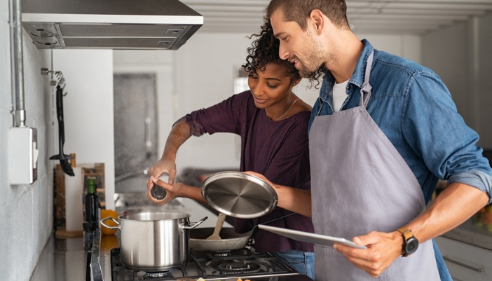 A couple stirs a pot on a stove together and adds salt. 