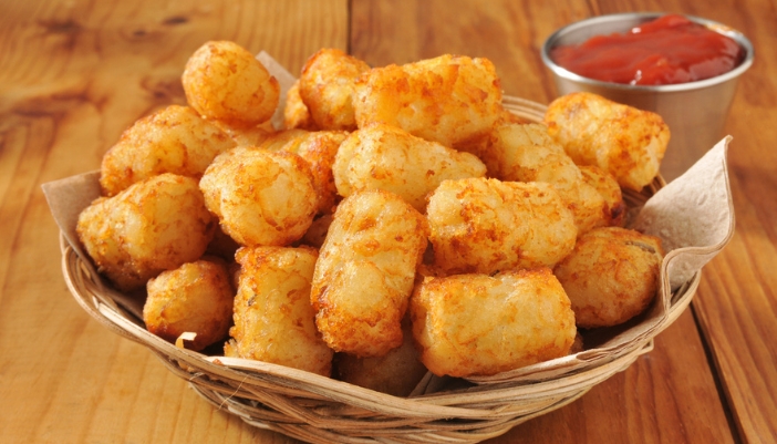 A basket of tater tots on a wooden surface with a dipping sauce on the side.