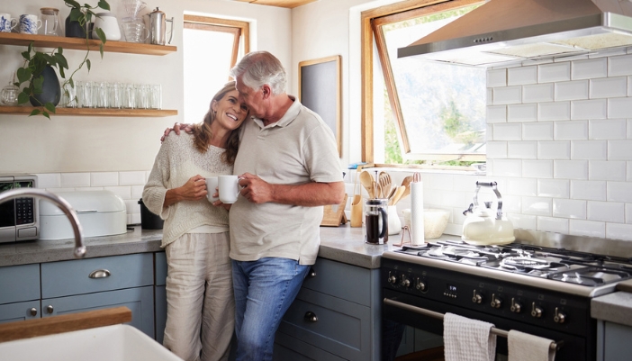 An elderly couple rests together in their home kitchen drinking coffee. 