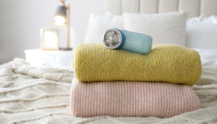 Two sweaters sit on a bed with a fabric shaver resting on top.