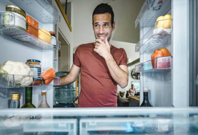 view from the inside of an open french door refrigerator of a man looking in wearily at the food inside