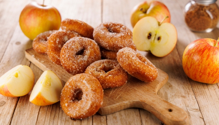 A wooden carrying tray of doughnuts surrounded by apples.