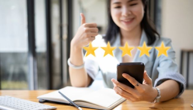 Front view of a woman smiling and looking at her phone with a thumbs up, and five yellow stars on top of the blurred image to indicate reviews.