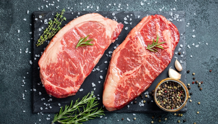 Raw steaks rest on a black slate cutting board with salt, pepper, and herbs