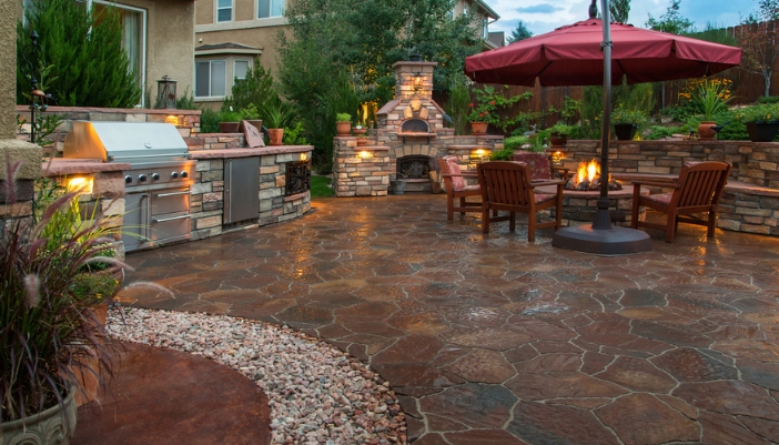 modern, luxurious backyard with an outdoor kitchen, grill, and lit firepit surrounded by chairs.