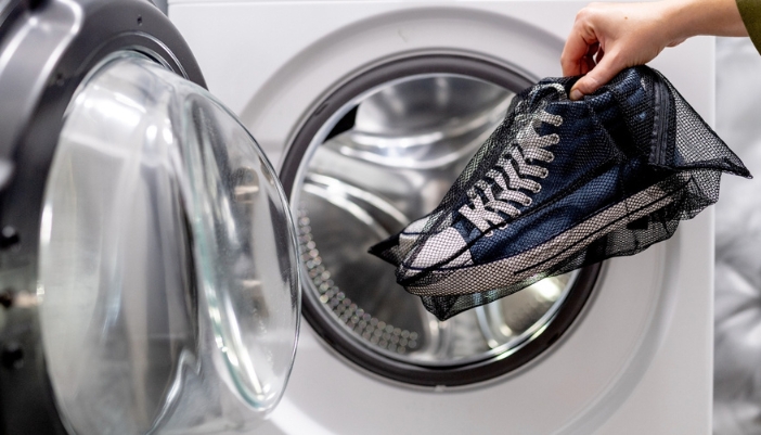 A hand places a pair of blue shoes in a mesh bag into a white dryer.