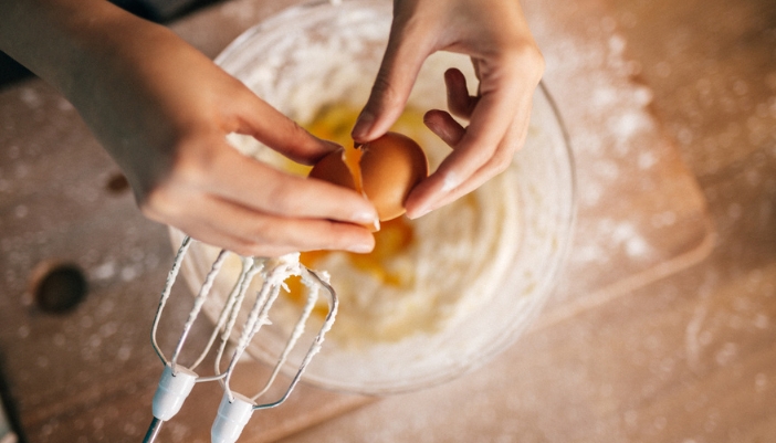 A pair of a hands cracks a brown egg over a bowl of cupcake batter.
