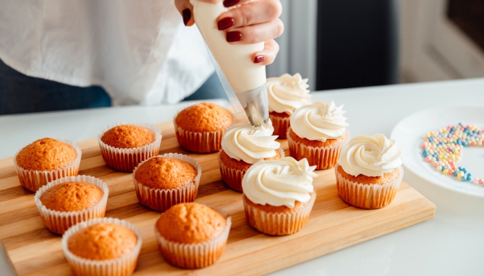 A pair of hands squeezes a frosting bag of white frosting on vanilla cupcakes on a wooden cutting board.