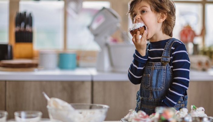 A young boy with long brown hair takes a big bite out of a freshly frosted cupcake.