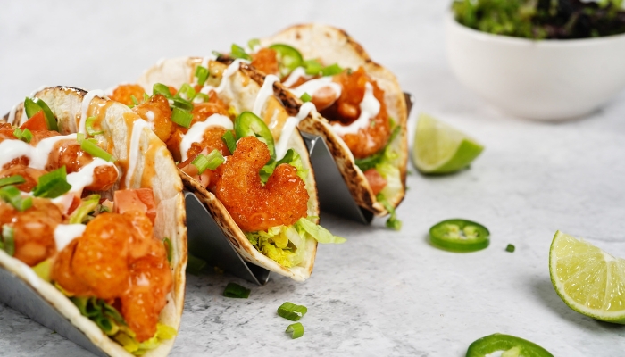 Three shrimp tacos are resting in a silver taco holder surrounded by limes and peppers.