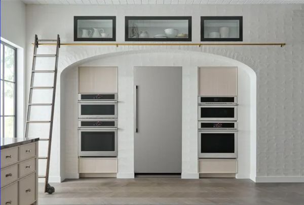 front view of an all white, luxury kitchen with a built in column refrigerator in the center wall, surrounded by built in wall ovens on both sides