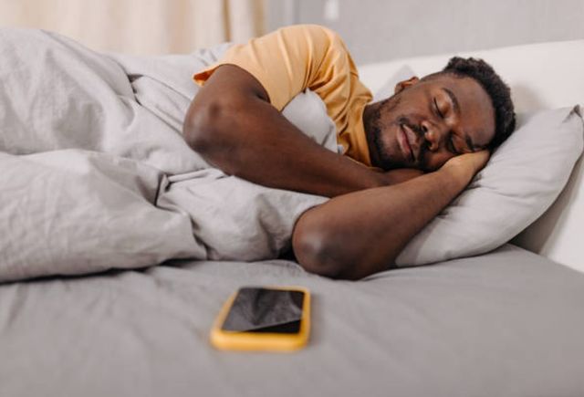 Side view of a man in an orange shirt sleeping in a bed of gray covers with his cell phone sitting on the bed in front of him