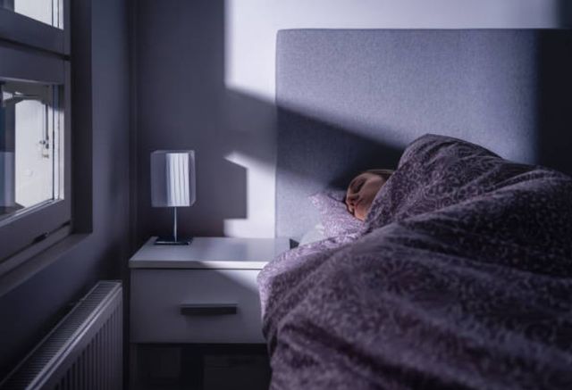 front view of a dim bedroom with light peeking in, and a person laying in bed covered by a blanker