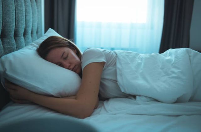 side view of a woman wearing a white shirt peacefully sleeping in a bed with white pillows and comforter and an upholstered headboard. 