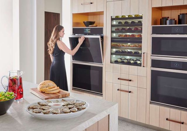 view of a woman opening the top oven of a double oven built into a wall beside a wine fridge and a second double oven, in the foreground is a white island with oysters and a breadboard