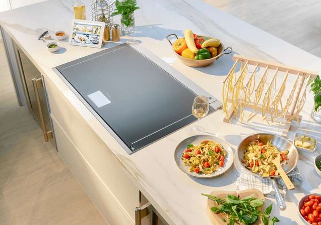 top view of a thermador induction range in a white marble countertop surrounded by cooking ingredients and a small tablet with a recipe on it and cooked pasta on the right