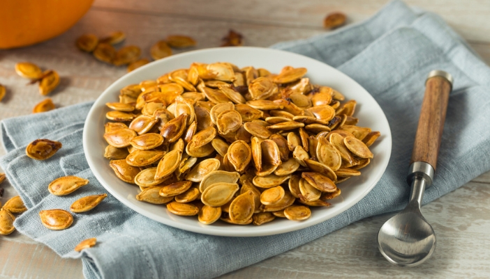 A bowl of roasted pumpkin seeds resting on a blue napkin with a spoon beside.