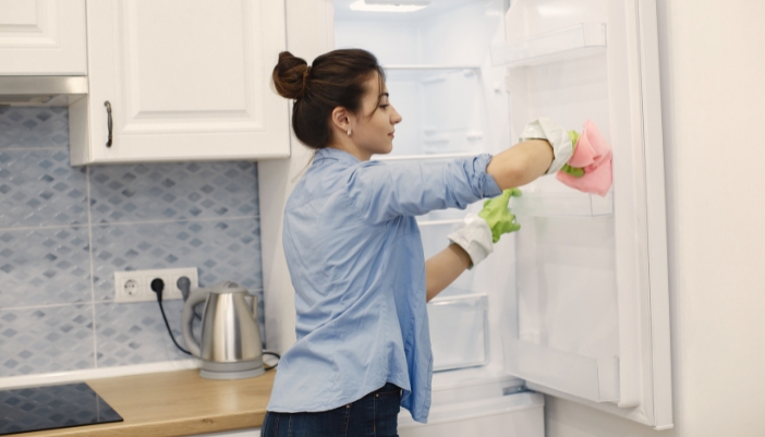 A young woman cleans out an empty fridge with a pink cloth.