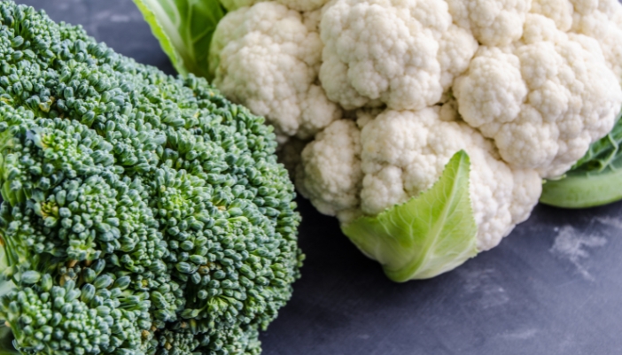 A head of broccoli and a head of cauliflower resting on a gray surface.