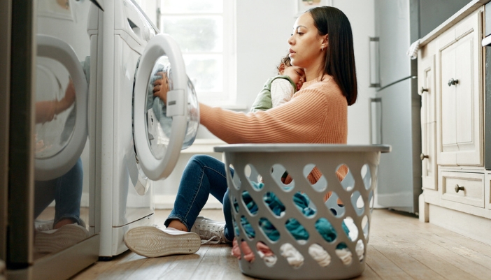 A woman holds a baby to her chest while she sits on the floor and loads her washer.